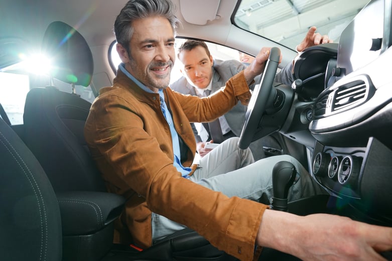 Man sitting in new car while salesman looks on behind him. 