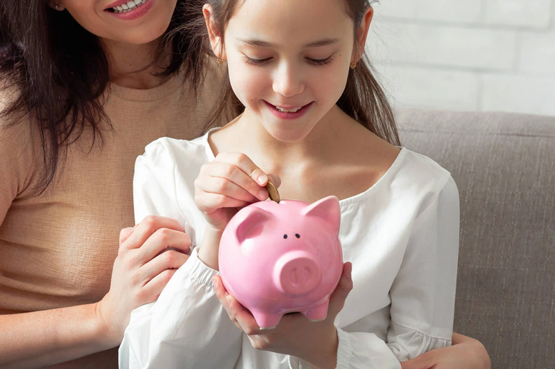Child placing money into a pink piggy bank with an adult guiding early savings habits at home