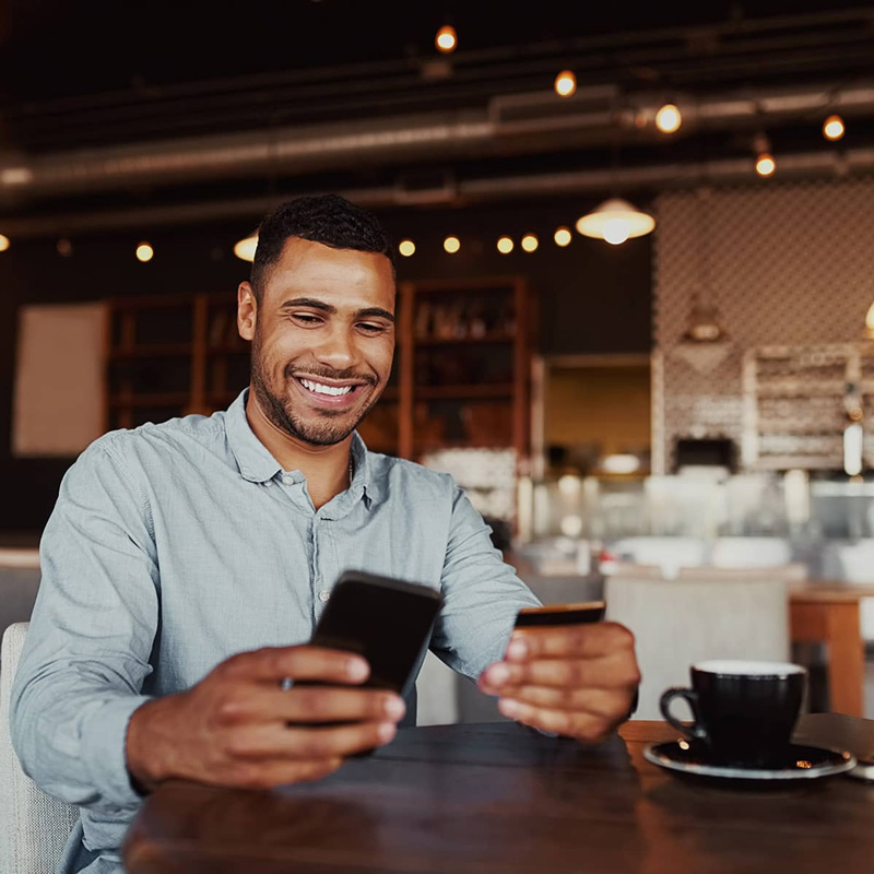 Customer sitting in a coffee shop, using a mobile phone and holding a bank card to manage his checking account.