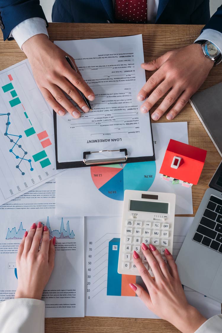 partial-view-of-customer-reading-loan-agreement-and-businesswoman-using-calculator-while-sitting-at-workplace-near-laptop,-house-model,-graph