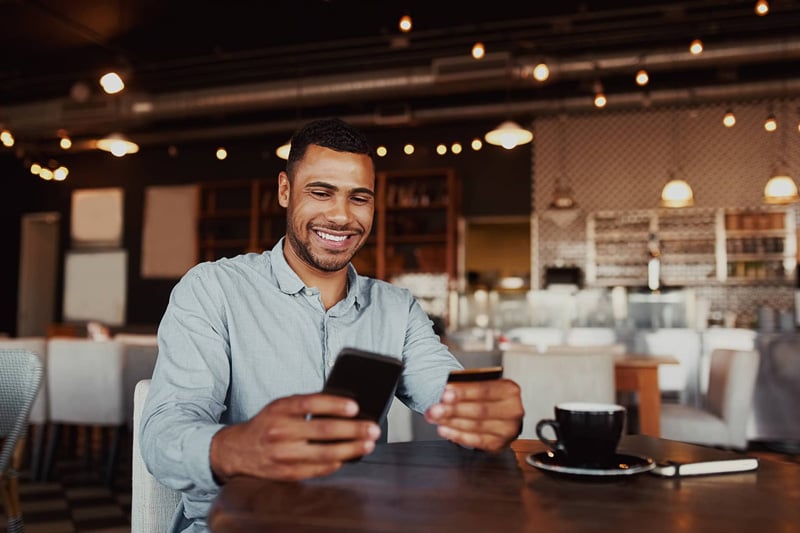 Person sitting at a cafe table using a smartphone and holding a debit card with a drink nearby.